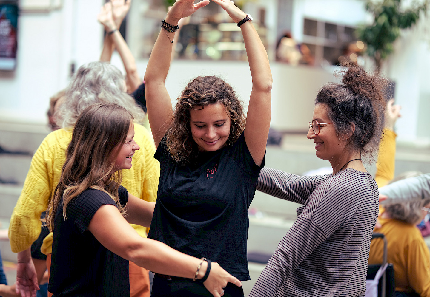 Drei Frauen tanzen. Eine Tänzerin in der Mitte hebt die Arme in die Luft, die beiden anderen fassen sich um sie herum an den Händen.
