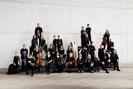 Die Musiker und Musikerinnen stehen als Gruppe mit ihren Instrumenten vor der Kamera