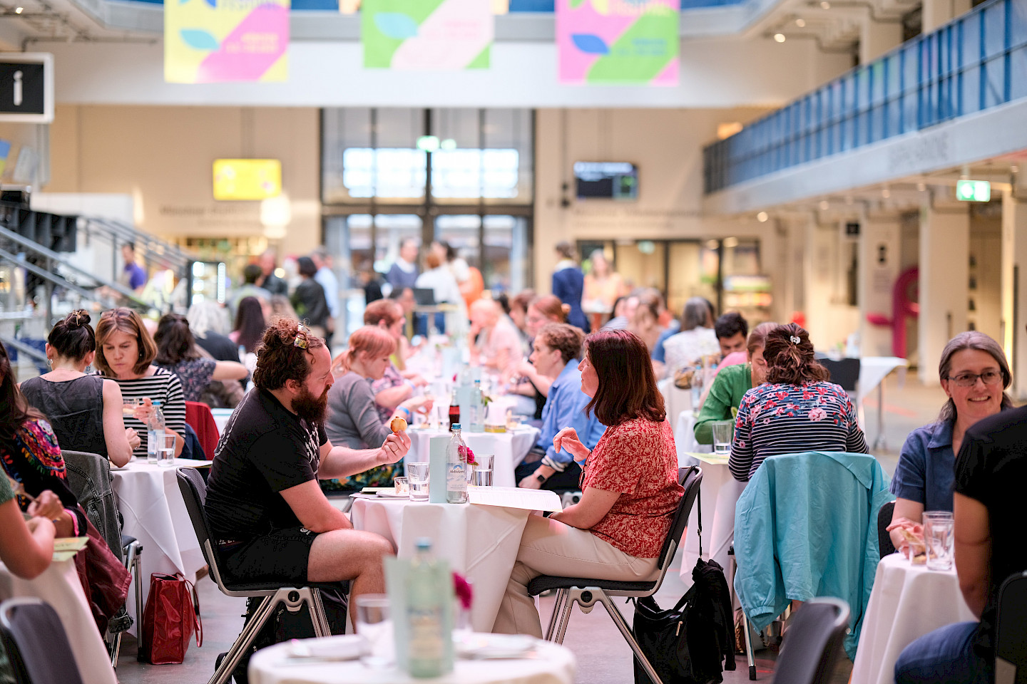 Menschen sitzen an Tischen in der Halle E und essen und reden miteinander.