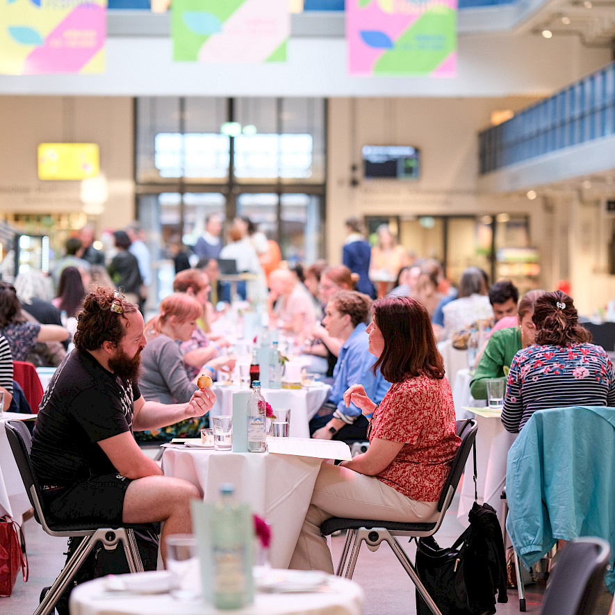 Menschen sitzen an Tischen in der Halle E und essen und reden miteinander. © Benedikt Feiten / Gasteig