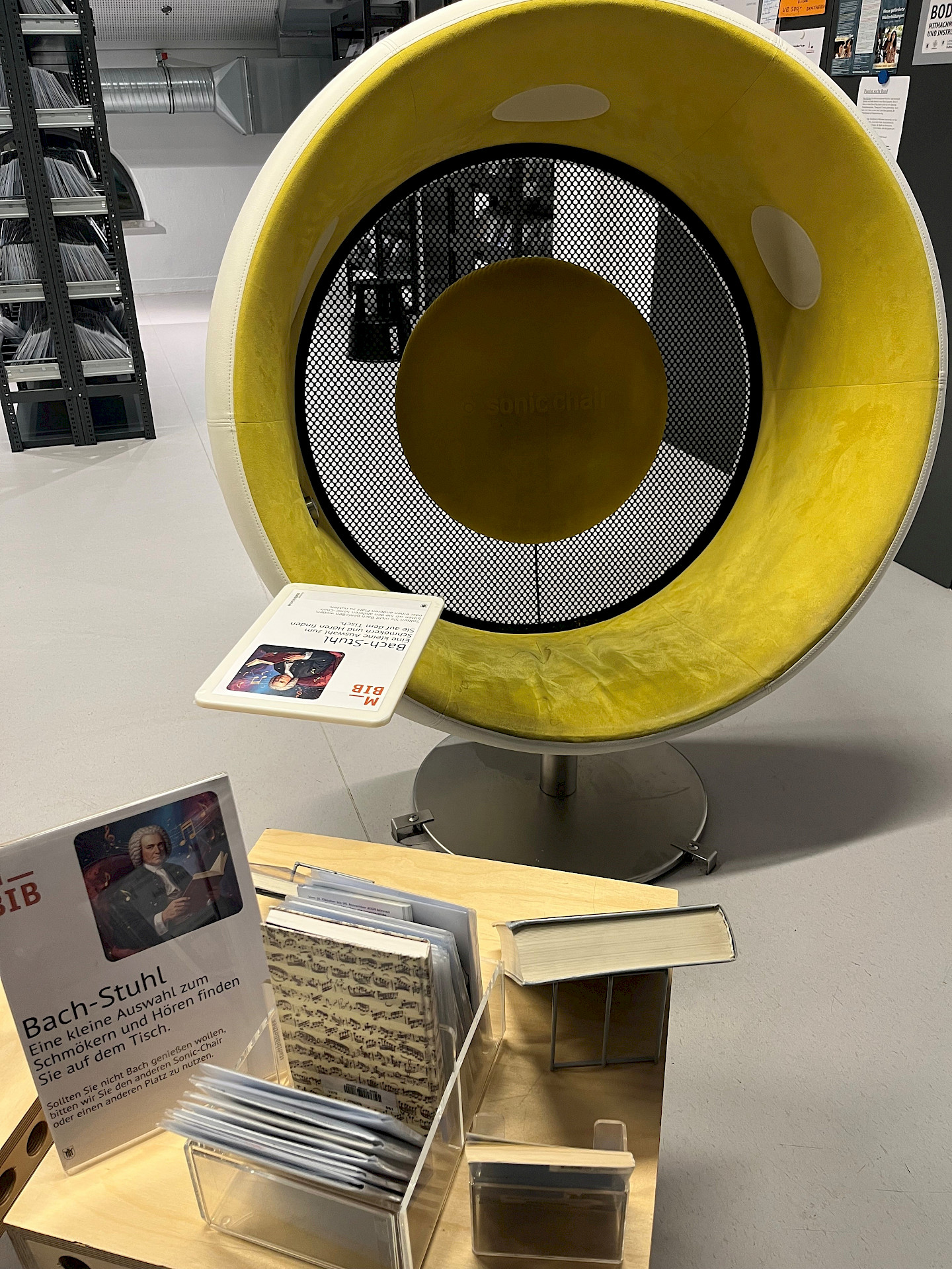 A round chair with a yellow cover, which you can sit in, stands behind a table with CDs and books on the subject of "Bach"