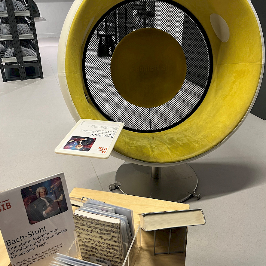 A round chair with a yellow cover, which you can sit in, stands behind a table with CDs and books on the subject of "Bach" © Münchner Stadtbibliothek