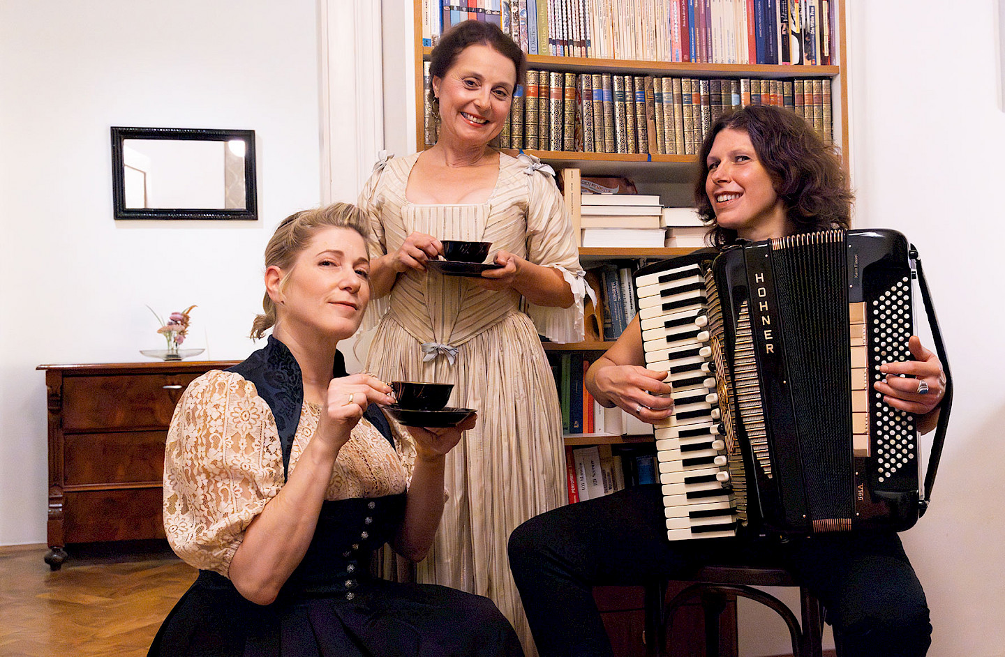 Links im Bild Ines Honsel als Anna Magdalena Bach. In der Mitte Gabi Altenbach als Christiana Mariana von Ziegler. Beide halten jeweils eine Kaffeetasse in der einen und eine Untertasse in der anderen Hand. Rechts auf dem Bild, auf einem Stuhl sitzend, Karin Füssel sie spielt auf dem Akkordeon.