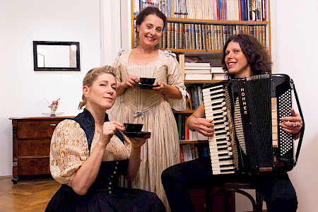 Links im Bild Ines Honsel als Anna Magdalena Bach. In der Mitte Gabi Altenbach als Christiana Mariana von Ziegler. Beide halten jeweils eine Kaffeetasse in der einen und eine Untertasse in der anderen Hand. Rechts auf dem Bild, auf einem Stuhl sitzend, Karin Füssel sie spielt auf dem Akkordeon.