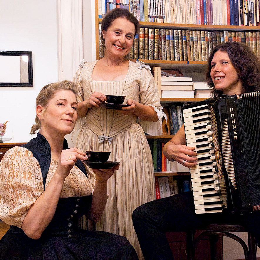 Links im Bild Ines Honsel als Anna Magdalena Bach. In der Mitte Gabi Altenbach als Christiana Mariana von Ziegler. Beide halten jeweils eine Kaffeetasse in der einen und eine Untertasse in der anderen Hand. Rechts auf dem Bild, auf einem Stuhl sitzend, Karin Füssel sie spielt auf dem Akkordeon. © HANS.cp