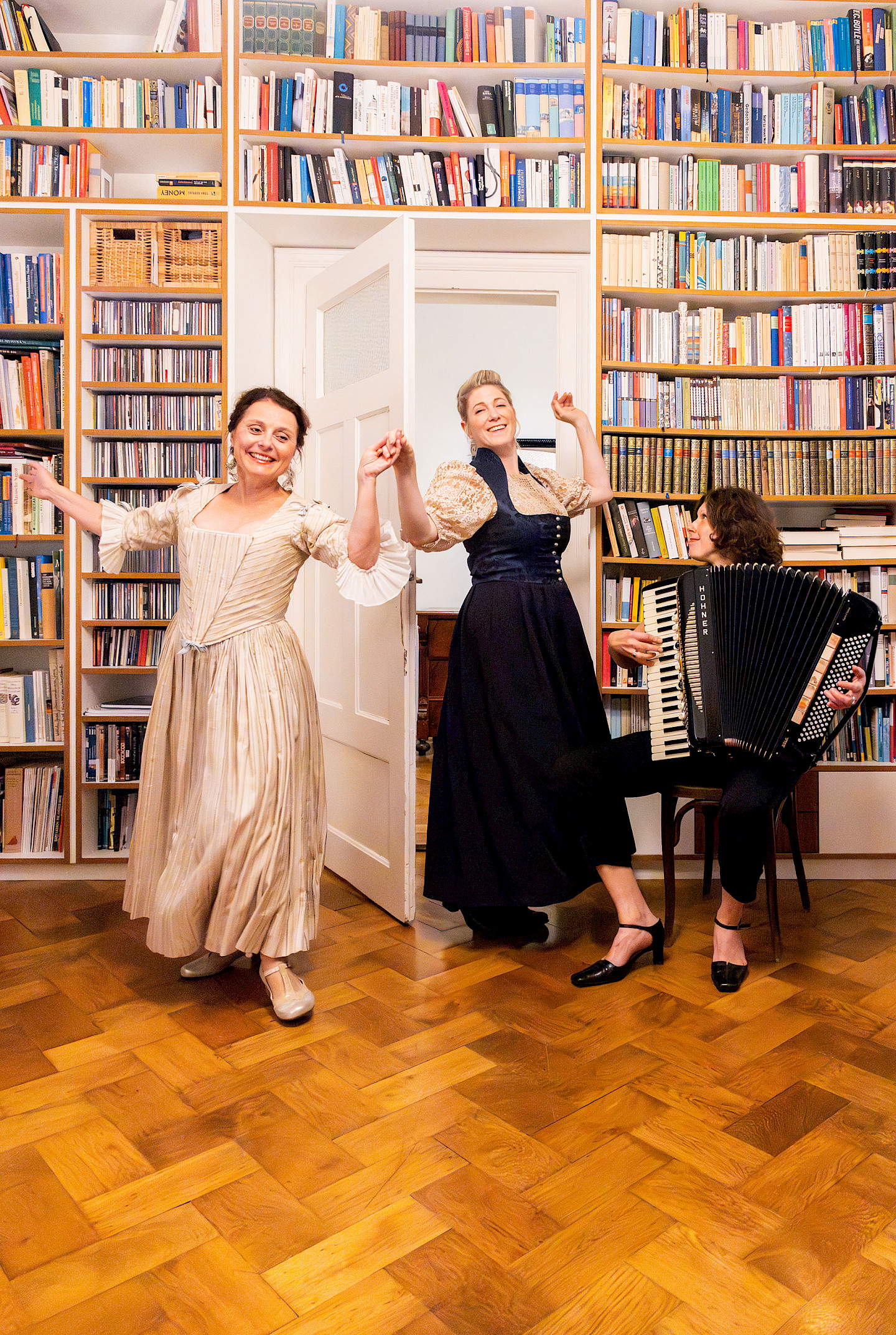 Links im Bild Gabi Altenbach als Christiana Mariana von Ziegler. Ines Honsel als Anna Magdalena Bach in der Mitte. Beide tanzen. Rechts auf dem Bild, auf einem Stuhl sitzend, Karin Füssel - sie spielt auf dem Akkordeon.